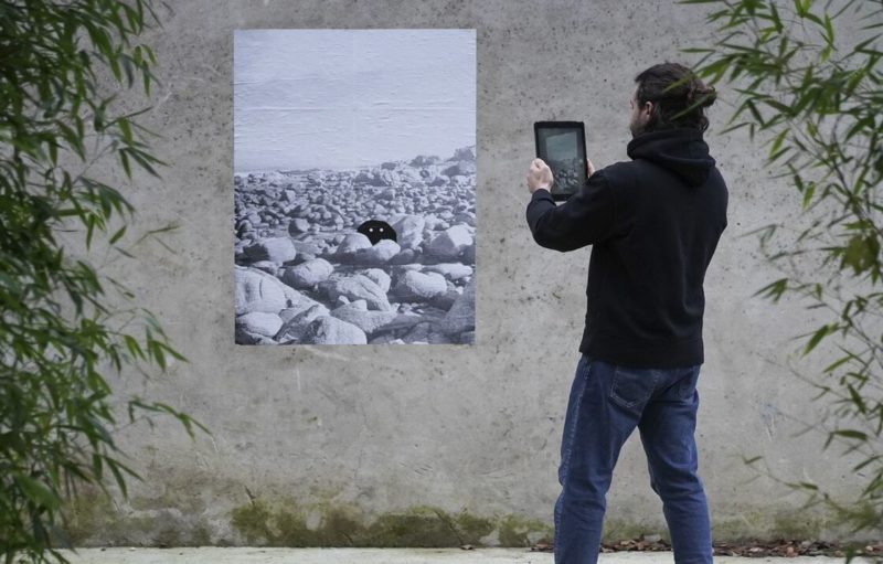 Photo d'un homme qui prend en photo une affiche de l'exposition en réalité augmentée Faune avec sa tablette numérique dans la rue.